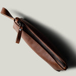 A brown leather zippered pouch with a matching leather wrist loop, shown diagonally on a light gray background with a soft shadow.