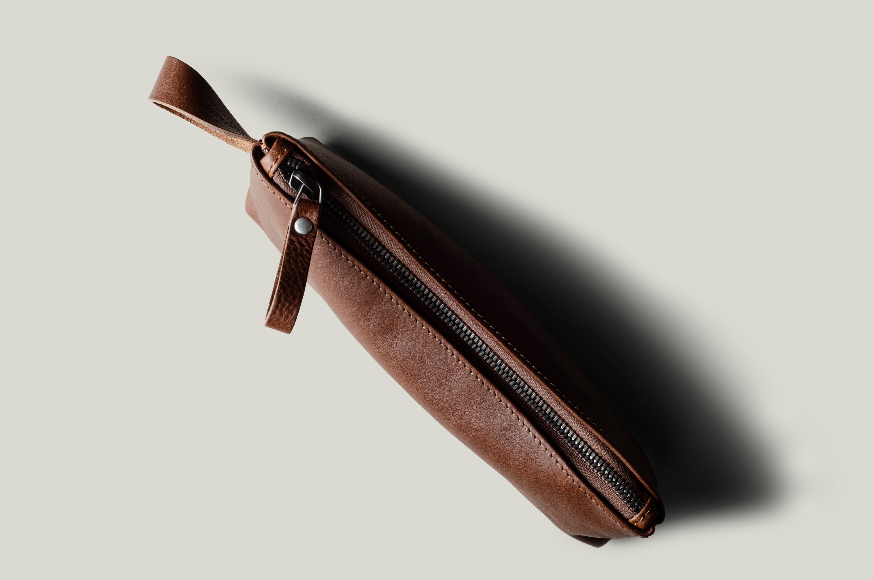 A brown leather zippered pouch with a matching leather wrist loop, shown diagonally on a light gray background with a soft shadow.