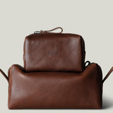 Two brown pebbled leather toiletry bags stacked together on a light gray background. The smaller rectangular zip pouch sits on top of a larger pouch.