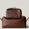 Two brown pebbled leather toiletry bags stacked together on a light gray background. The smaller rectangular zip pouch sits on top of a larger pouch.