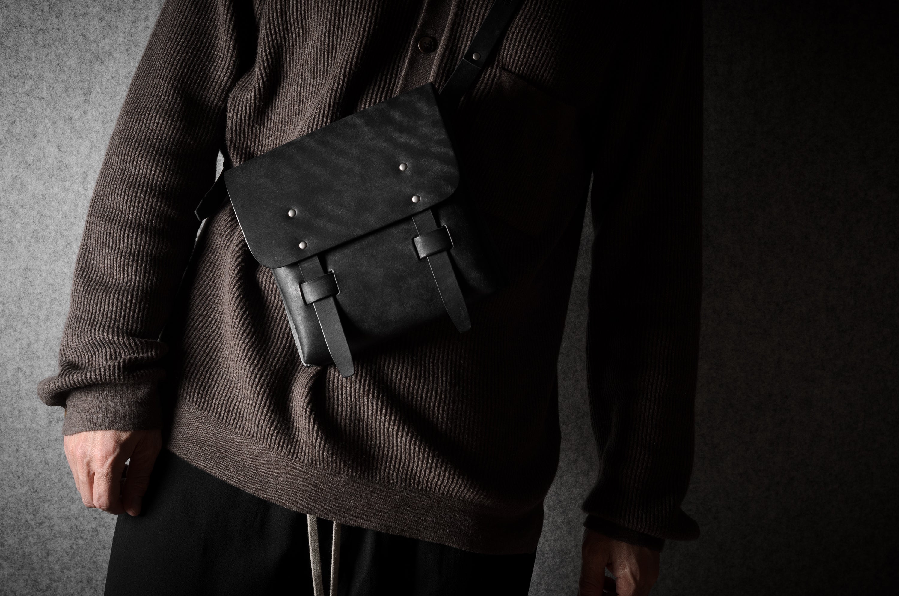 Person wearing a dark brown sweater with a black leather bag over one shoulder against a gray background