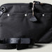 A charcoal-colored laptop bag with black leather straps and a brown wool-blend lining. It features a main compartment closed with a zipper and an additional exterior pocket.