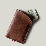 A brown textured leather card holder wallet with a small red tab, shown open with folded dollar bills partially sticking out, on a light gray background.