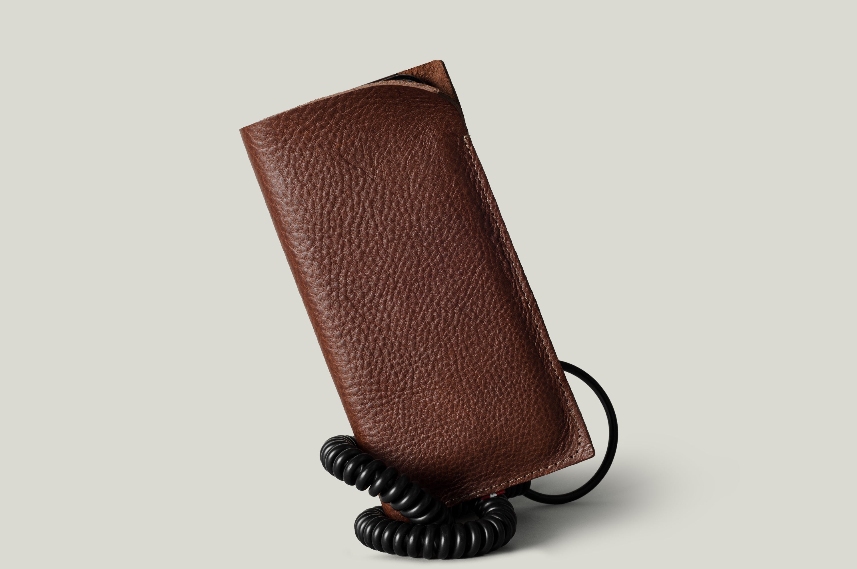 A brown textured leather iPhone 18 Pro case with a black coiled cord, shown standing upright on a light gray background.