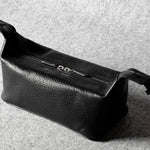 A black textured leather toiletry pouch with a top zipper and adjustable strap, photographed on a gray felt surface.