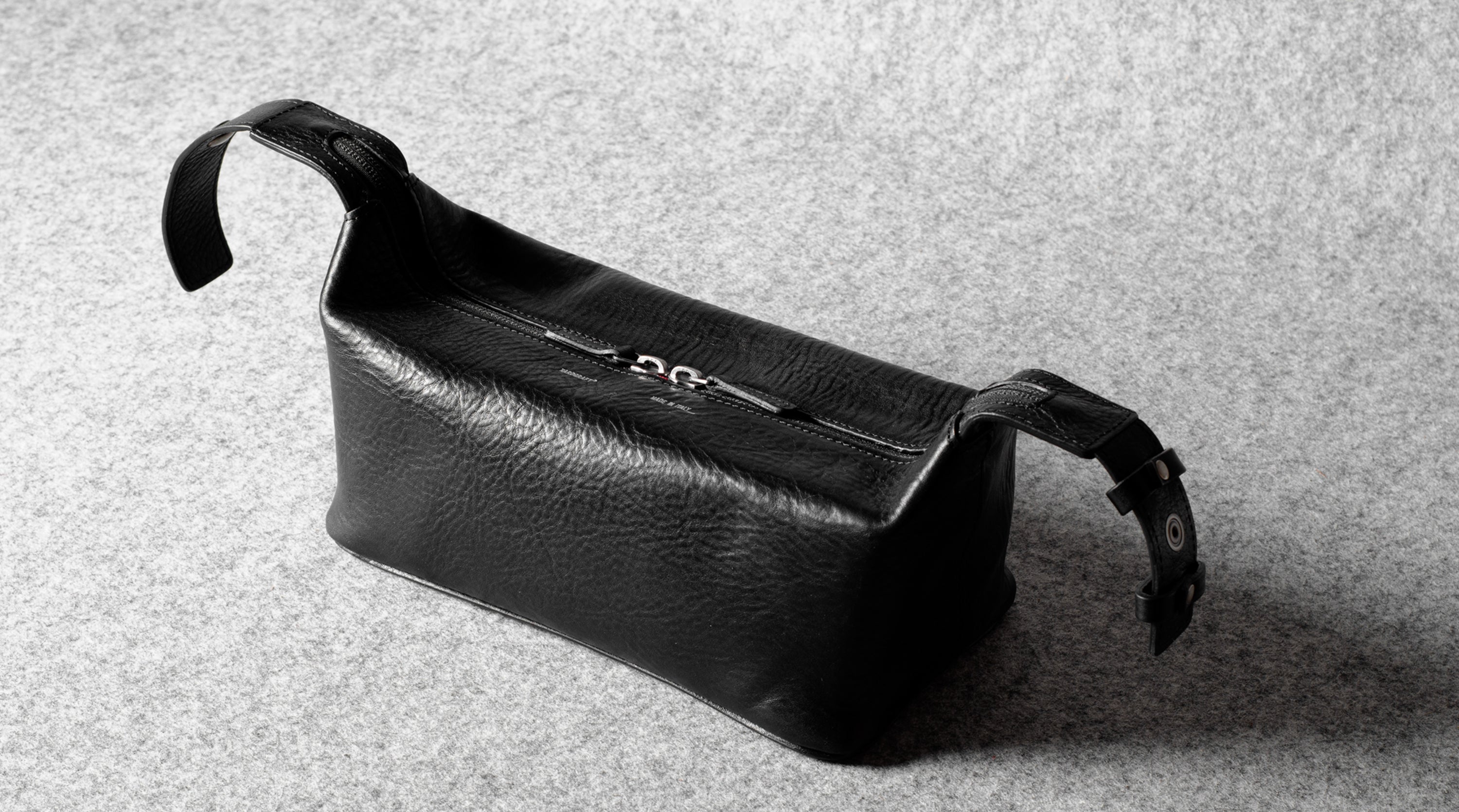 A black textured leather toiletry pouch with a top zipper and adjustable strap, photographed on a gray felt surface.