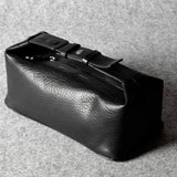 A black textured leather toiletry pouch with a buckle strap closure, photographed on a gray felt surface.