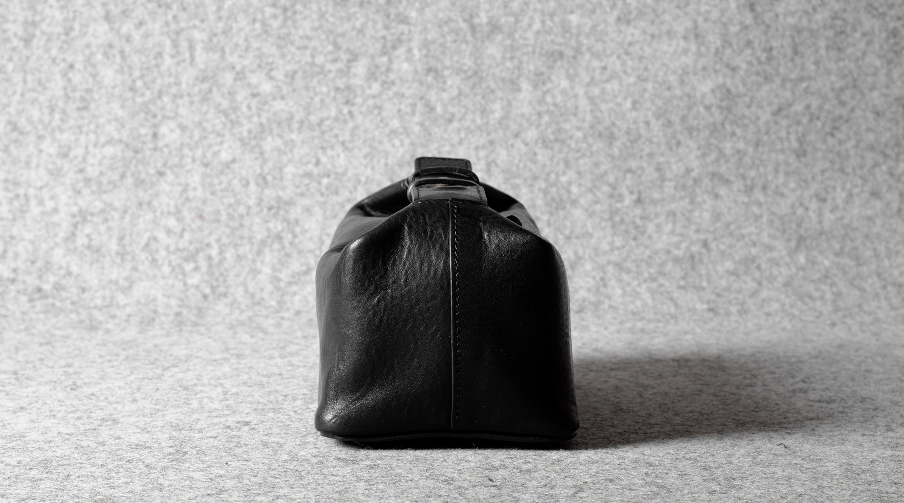 A black leather toiletry bag with a structured, rounded rectangular shape and a top handle, photographed from the back against a textured gray background.
