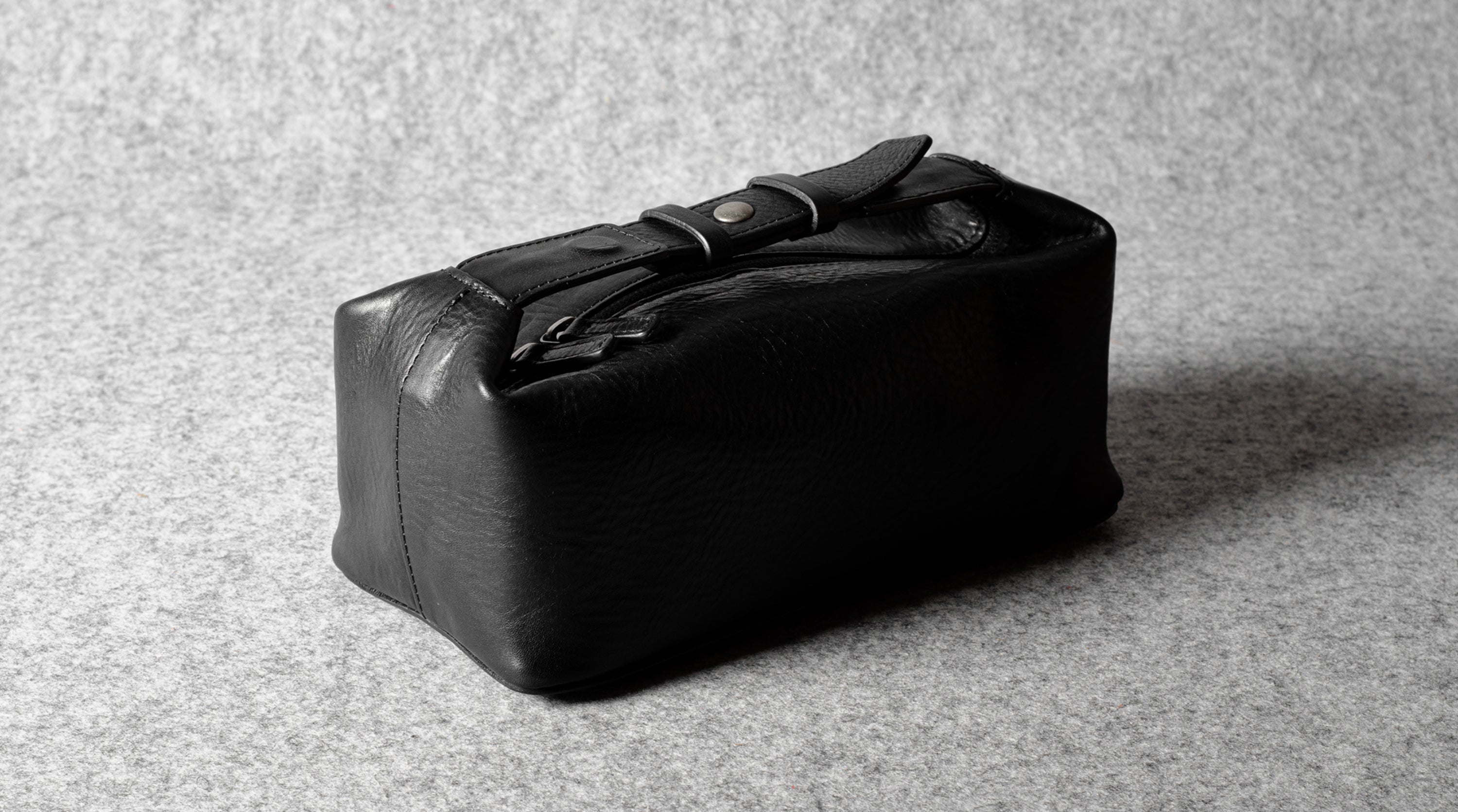 A black leather toiletry case with a strap and buckle closure, photographed on a textured gray surface.