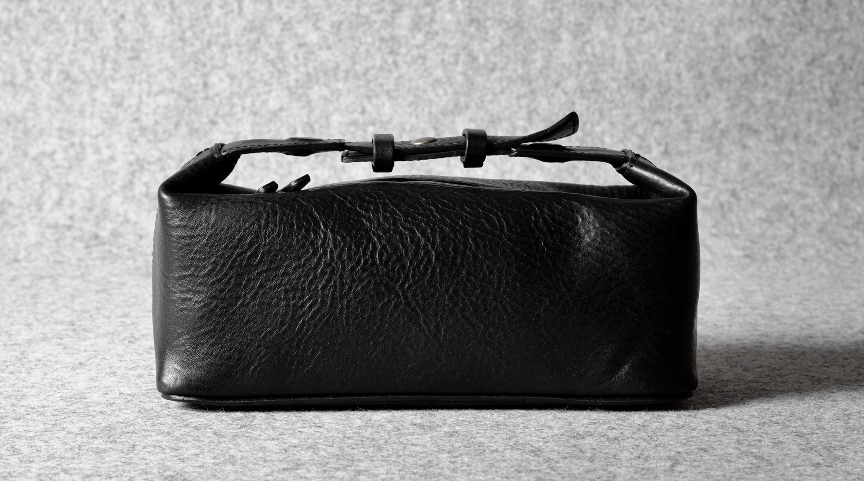 A black textured leather toiletry case with a top zipper and a thin strap detail, photographed on a neutral gray textured background.