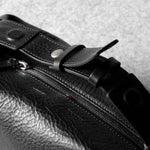 Close-up of a black textured leather toiletry bag with a zipper, snap-button strap, and metal hardware, photographed on a light gray surface.