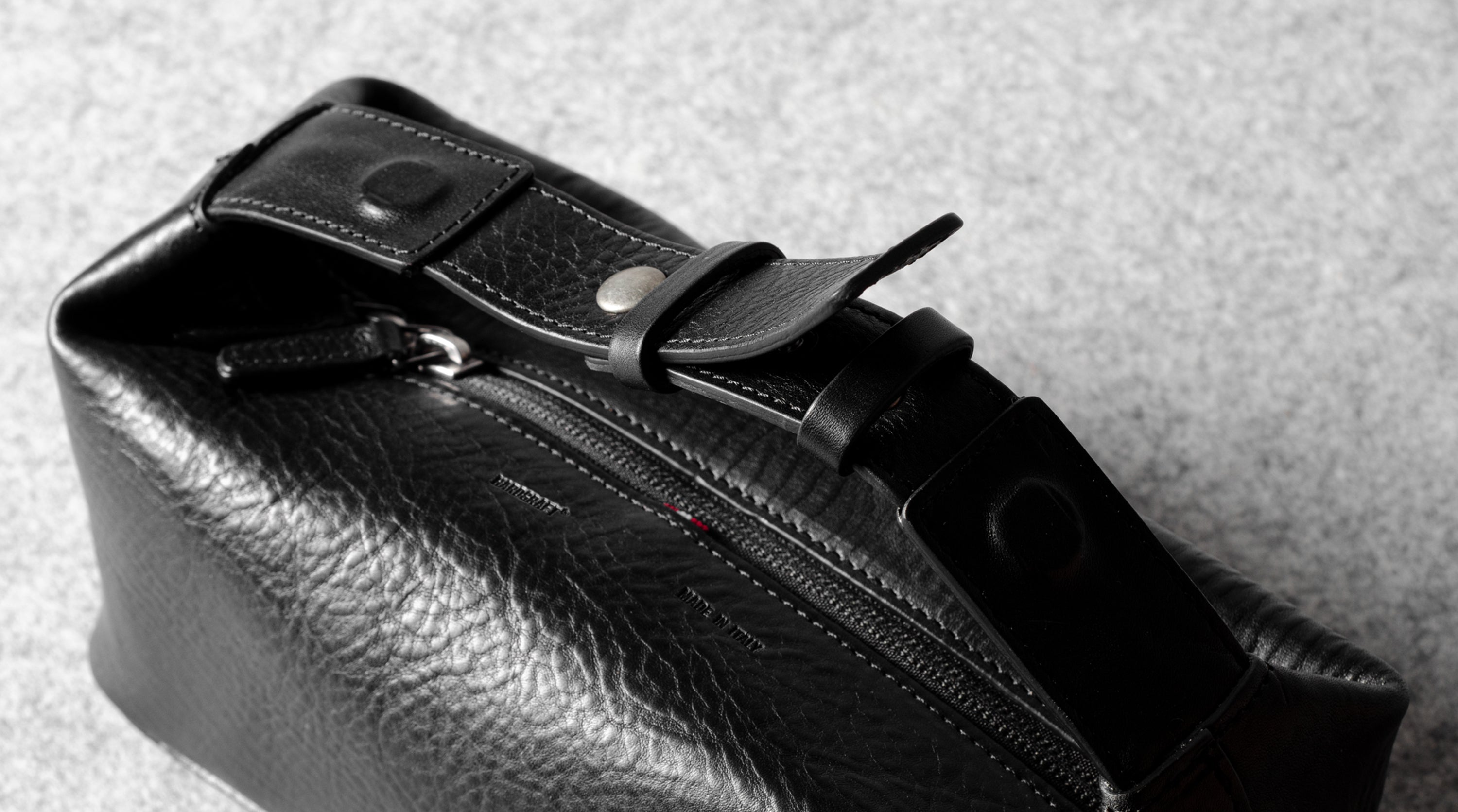 Close-up of a black textured leather toiletry bag with a zipper, snap-button strap, and metal hardware, photographed on a light gray surface.