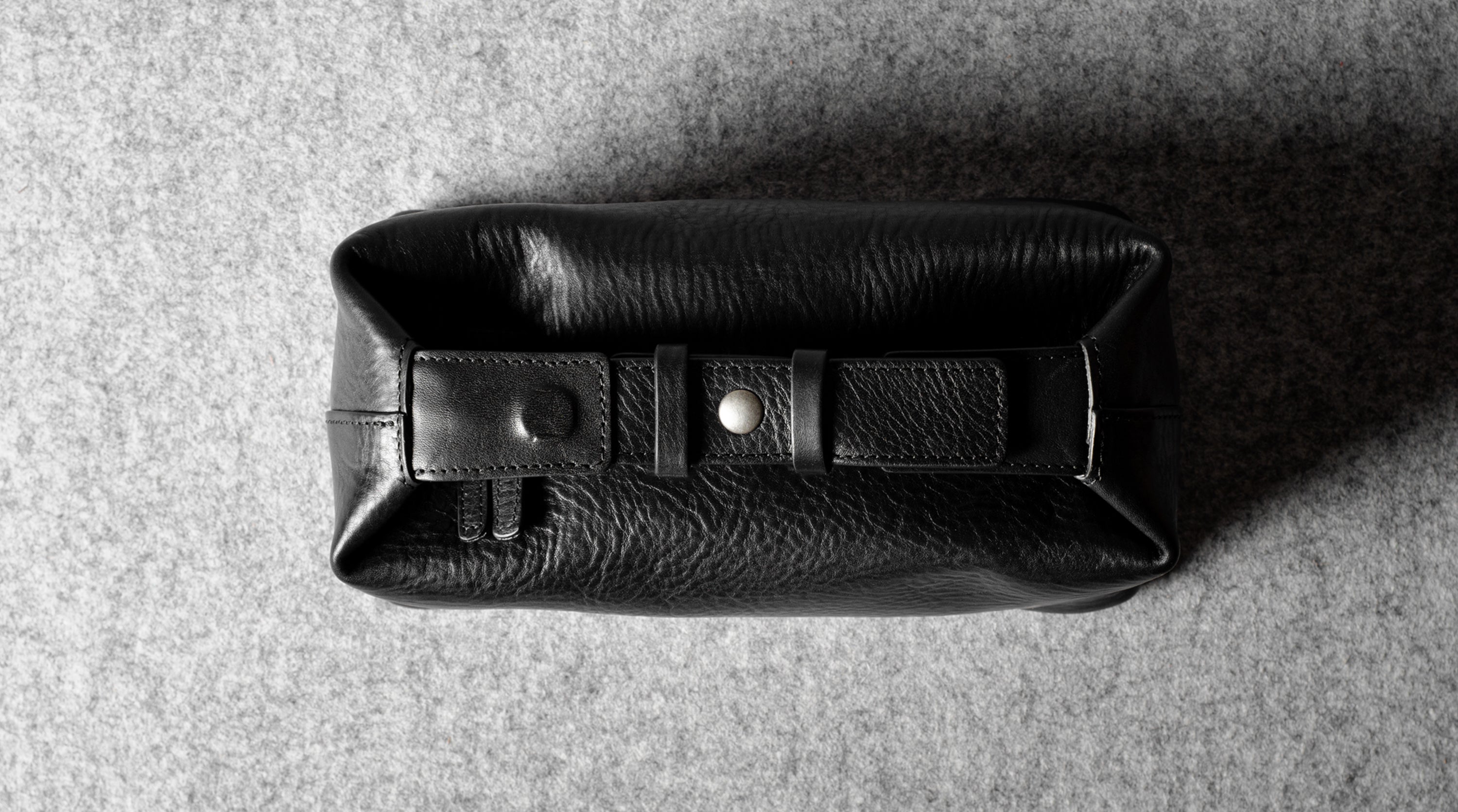 A black pebbled leather toiletry pouch with a buckle strap closure, photographed from a top-down angle on a textured gray background.