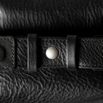 A close-up of a black leather belt with a silver-toned snap button and textured grain, laid horizontally across a matching black leather surface.