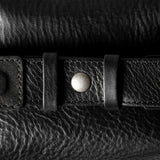 A close-up of a black leather belt with a silver-toned snap button and textured grain, laid horizontally across a matching black leather surface.