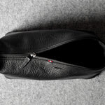 A black textured leather zippered toiletry pouch or small bag, shown from a top-down view on a gray felt surface.