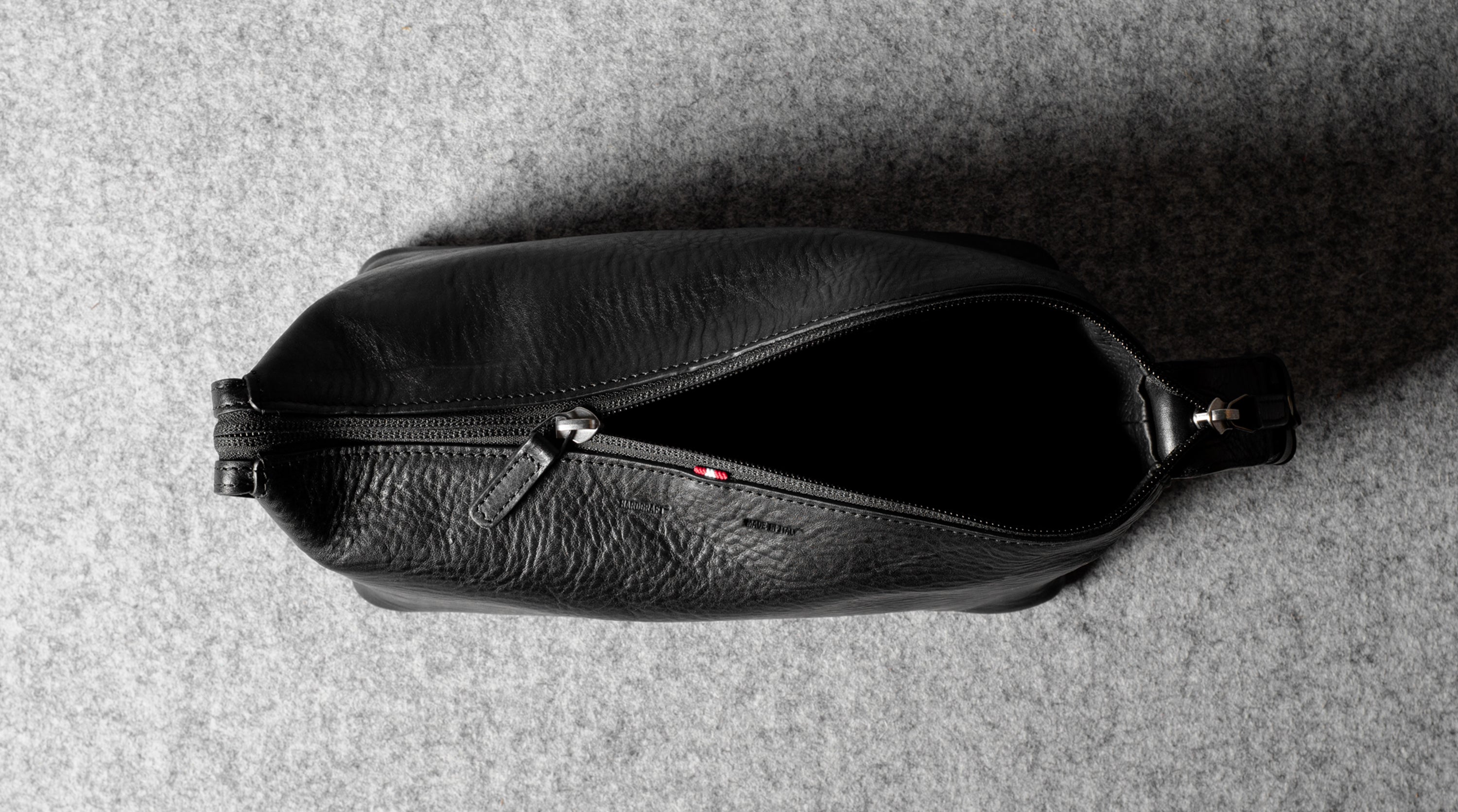 A black textured leather zippered toiletry pouch or small bag, shown from a top-down view on a gray felt surface.