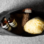 An open black zippered toiletry pouch on a gray textured background, containing two small amber bottles with black caps and white labels, a brown scrubber, and a yellow sponge.