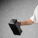 A person wearing a white t-shirt holds a black rectangular toiletry bag against a textured gray background.