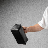 A person wearing a white t-shirt holds a black rectangular toiletry bag against a textured gray background.