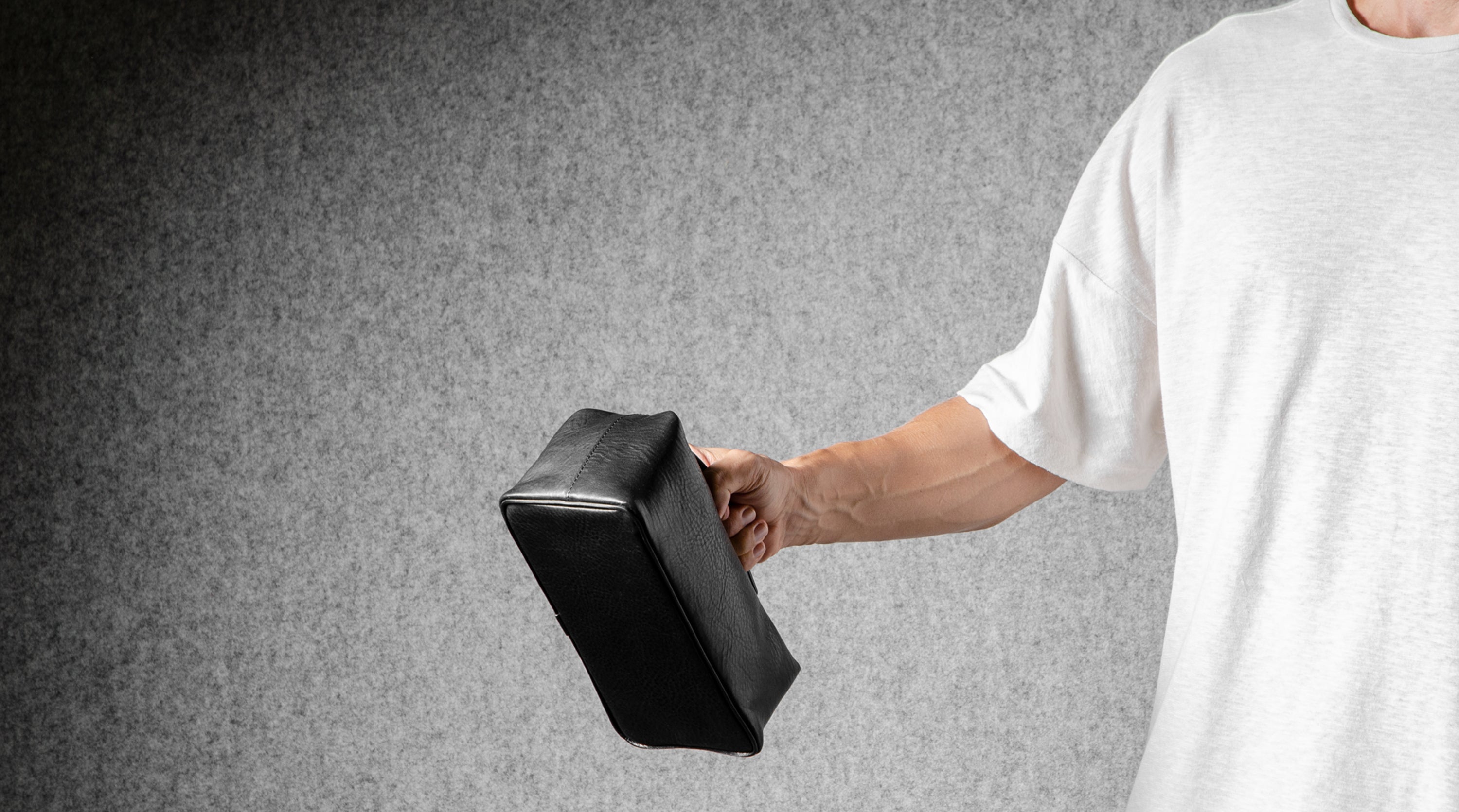 A person wearing a white t-shirt holds a black rectangular toiletry bag against a textured gray background.