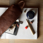 A brown leather zip pouch laid diagonally across an open notebook, surrounded by stationery items including a fountain pen, ink bottle, and small accessories on a wood tabletop.