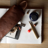A brown leather zip pouch laid diagonally across an open notebook, surrounded by stationery items including a fountain pen, ink bottle, and small accessories on a wood tabletop.
