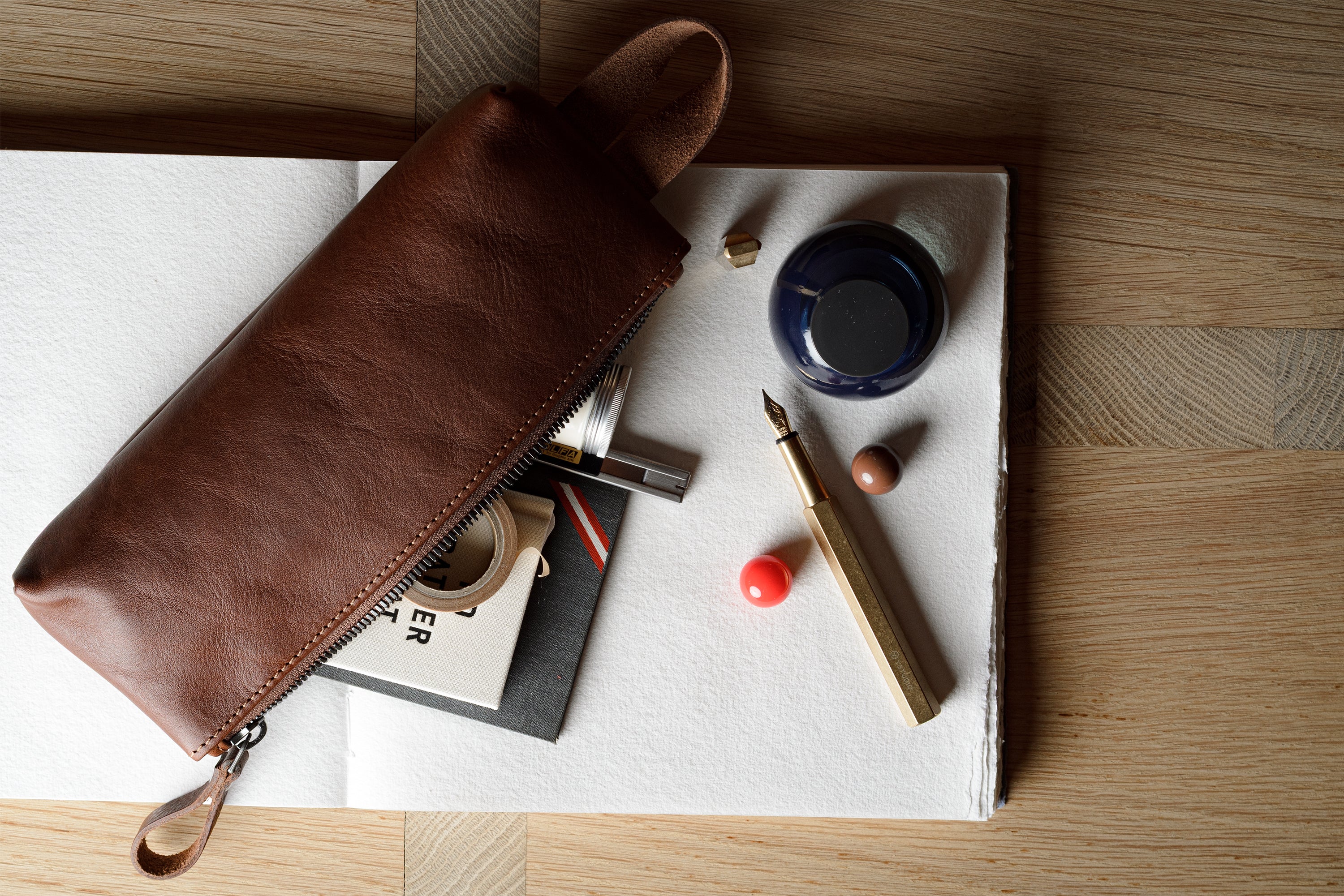 A brown leather zip pouch laid diagonally across an open notebook, surrounded by stationery items including a fountain pen, ink bottle, and small accessories on a wood tabletop.