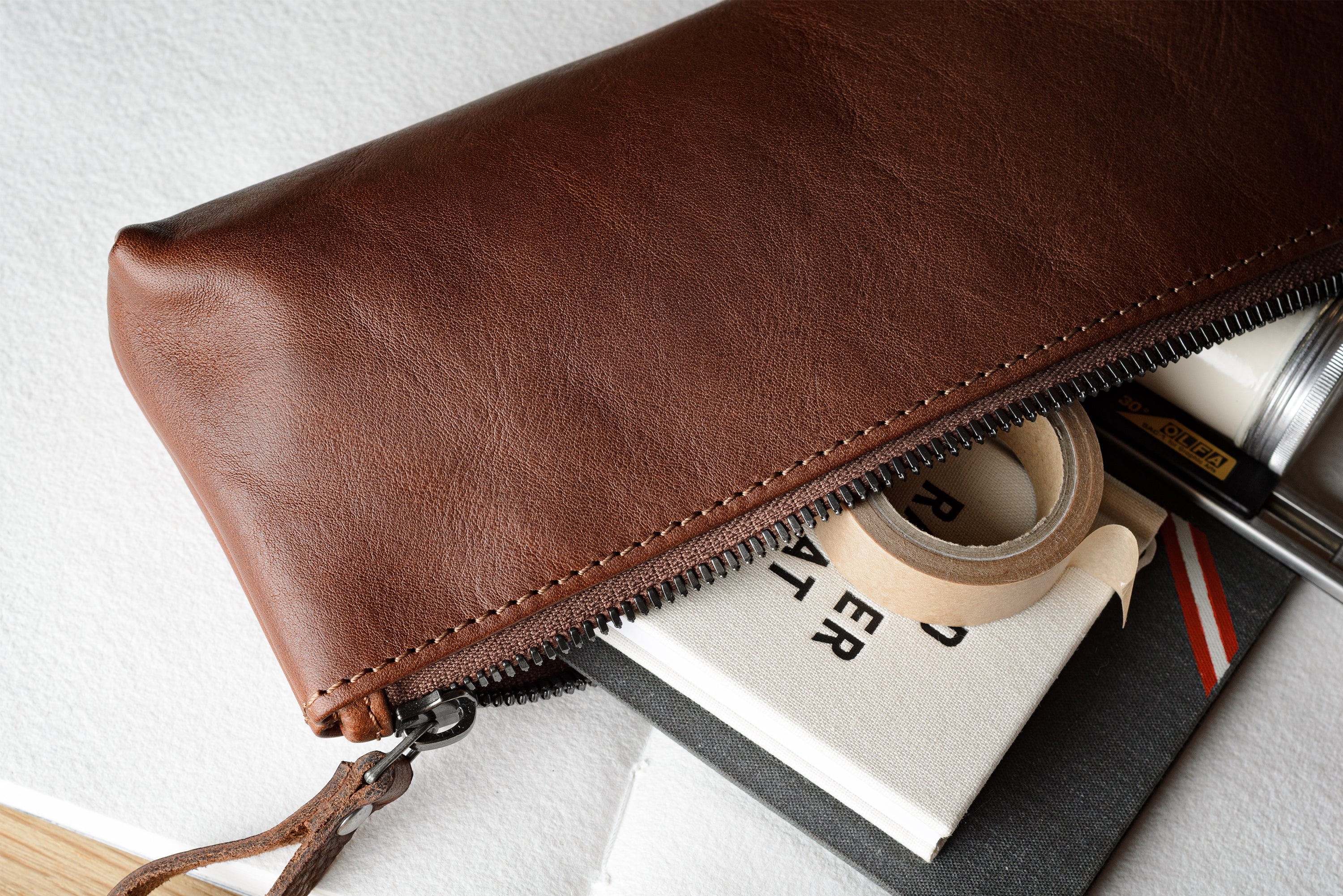 A brown leather zip pouch with a wrist strap, shown partially open and filled with stationery items like a notebook, tape, and pen, on a light textured background.