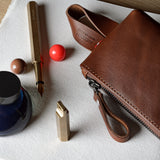 A brown leather zip pouch with a wrist strap, arranged on a white cloth alongside a gold pen, a blue ink bottle, a small red ball, and a gold lighter on a light wood surface.