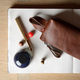 A brown leather zip pouch with a wrist strap, styled on a white sheet of paper over a light wood background alongside a gold pen, a small blue ink bottle, and decorative beads.