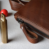 A close-up of a brown leather zip pouch with a wrist strap, shown on a light textured surface alongside stationery items including a fountain pen, gold-colored tools, and a red spherical object.