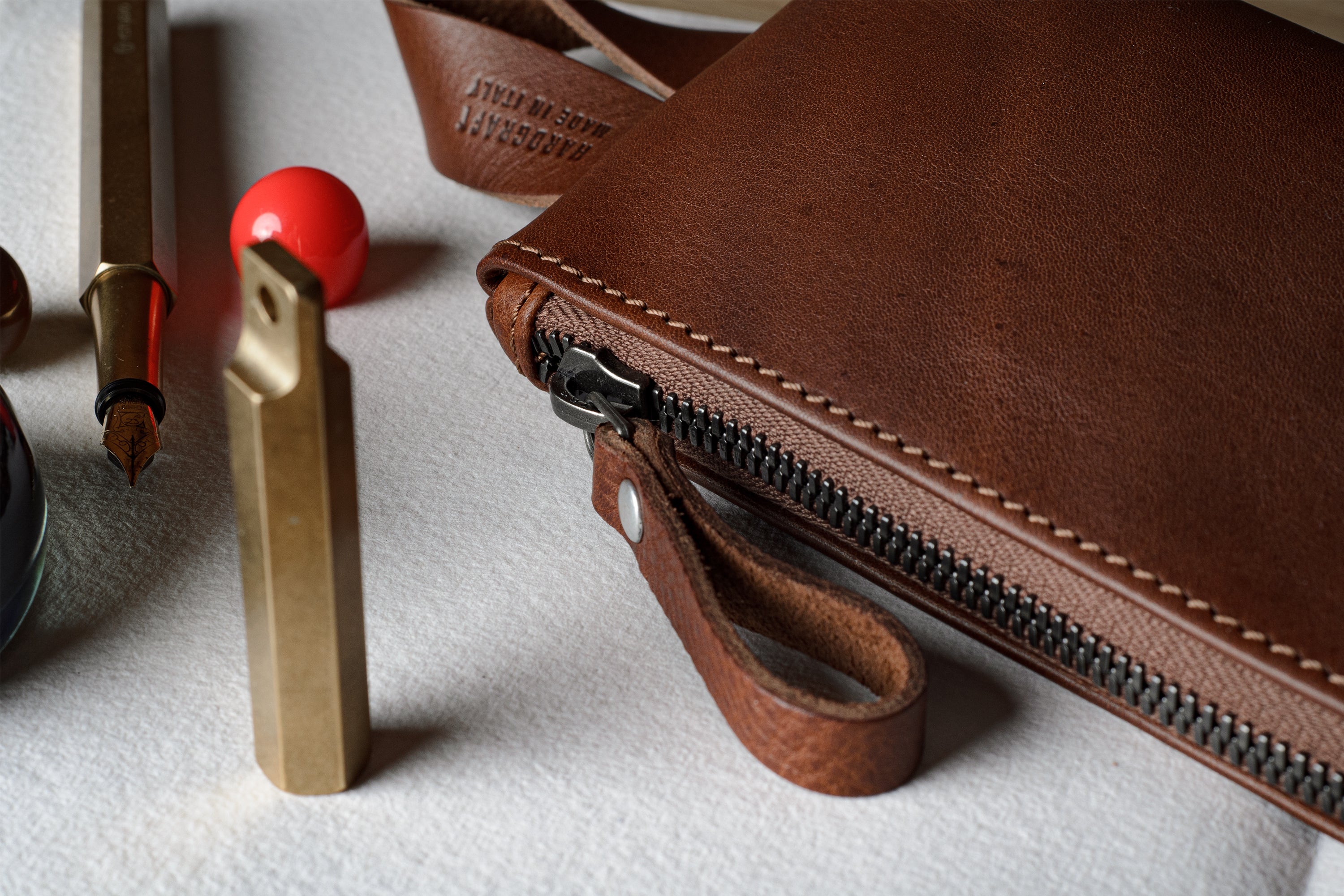 A close-up of a brown leather zip pouch with a wrist strap, shown on a light textured surface alongside stationery items including a fountain pen, gold-colored tools, and a red spherical object.