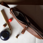 A brown leather zippered pencil case or pouch lies open on a white sheet of paper, surrounded by stationery props including a gold pen, ink bottle, and small red and brown objects, on a light wood surface.