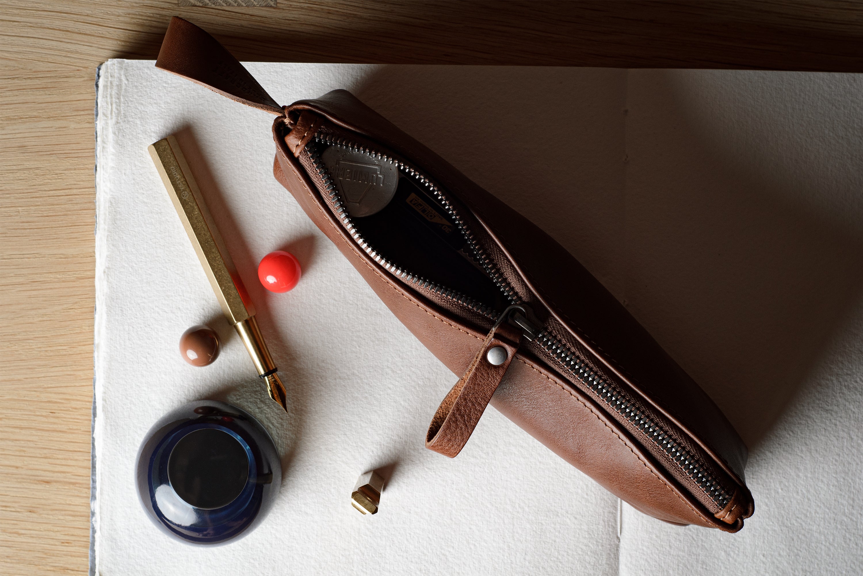 A brown leather zippered pencil case or pouch lies open on a white sheet of paper, surrounded by stationery props including a gold pen, ink bottle, and small red and brown objects, on a light wood surface.