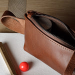 A close-up of a brown leather zippered pouch or cosmetic bag lying on a white cloth, with a gold pen and a small red sphere nearby on a wooden surface.