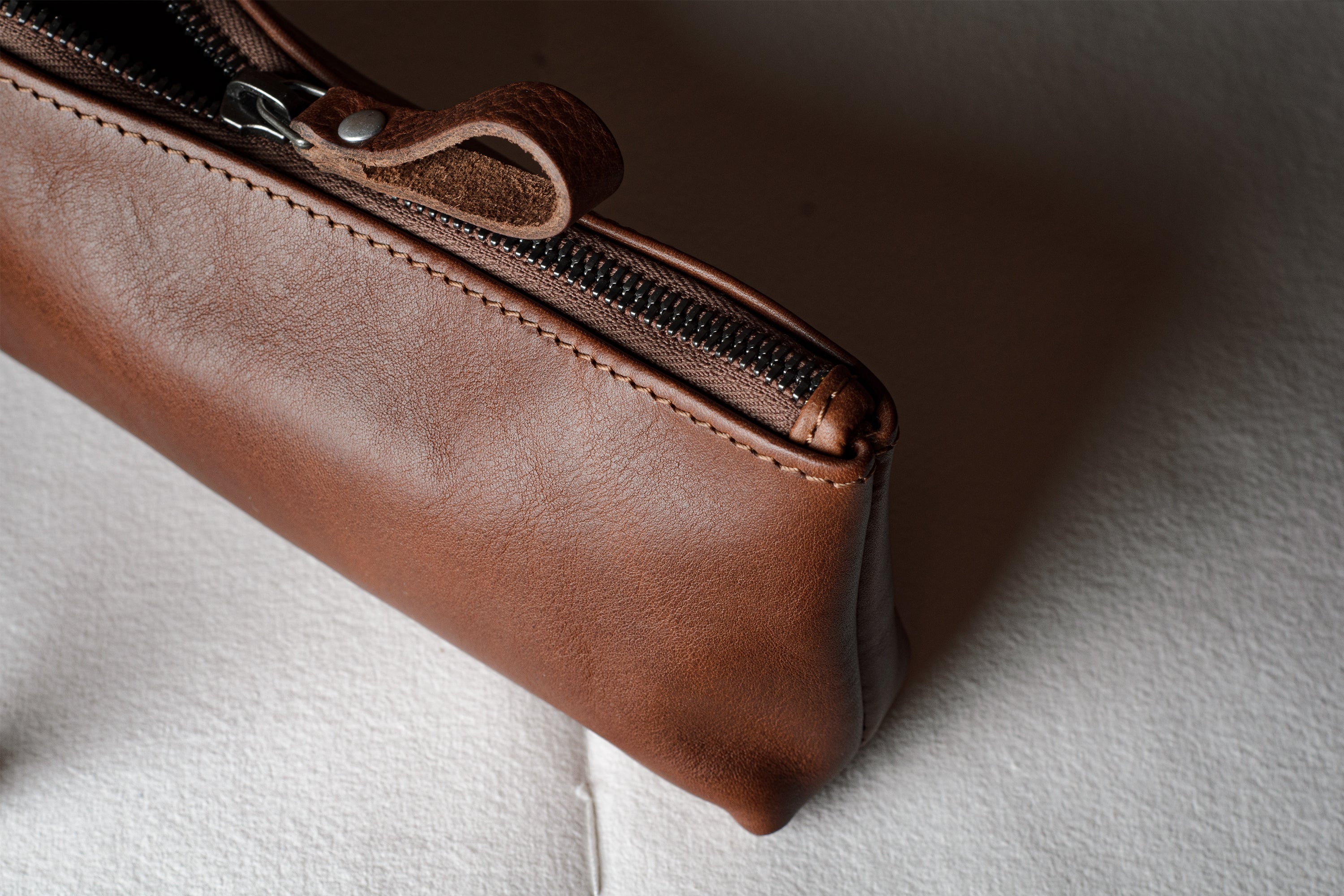 A close-up of a brown leather zippered pouch or case resting on a textured white fabric surface.