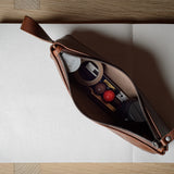 A brown leather zippered pouch with a pull tab, shown open on a white notebook over a light wood surface. The pouch contains various small cosmetic items.