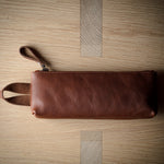 A brown leather zip pouch with a wrist strap, laid flat on a light wood grain surface.