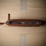 A brown leather zippered pouch with a leather pull tab, laid horizontally on a light wood grain background.