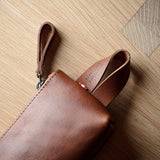 A close-up of a brown leather pouch or sleeve with a wrist strap, placed on a light wood floor.