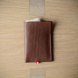 A brown textured leather card holder wallet with visible stitching and a small red-and-white cord detail, photographed from above on a light wood grain surface.
