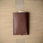 A brown textured leather card holder wallet with a small red and white striped ribbon bookmark, displayed on a light wood grain surface.