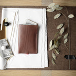 A brown leather card holder wallet with a small red tab, displayed on a brown leather folder with white paper, a gold clip, and dried leaves on a wooden table.