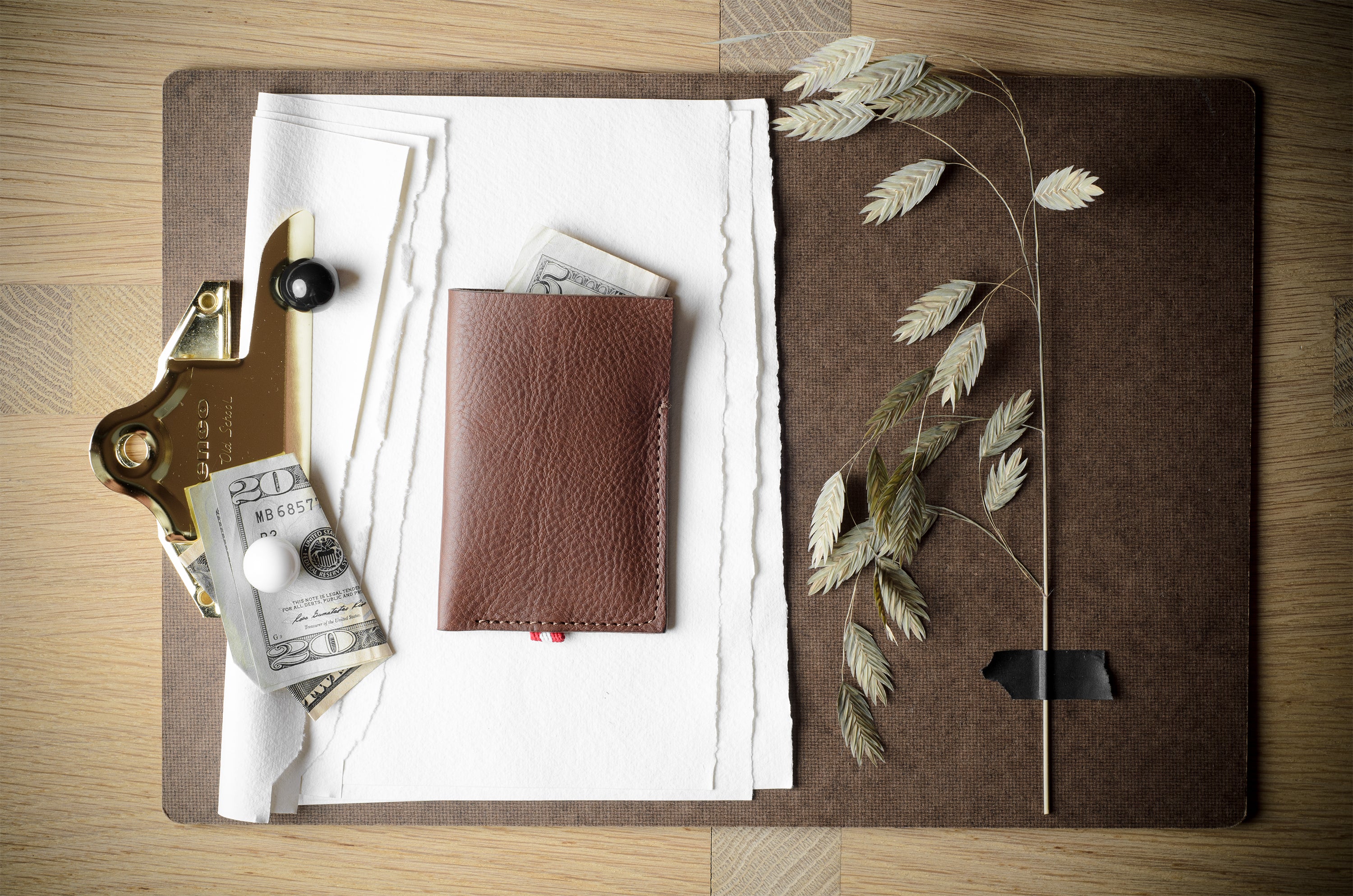 A brown leather card holder wallet with a small red tab, displayed on a brown leather folder with white paper, a gold clip, and dried leaves on a wooden table.