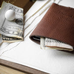 A brown leather card holder or slim wallet with a few dollar bills and a small white marble inside, styled on a white textured paper surface with a wooden table edge visible.