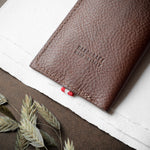 A brown textured leather notebook cover with 'HARDGRAFT MADE IN ITALY' debossed on the front, styled on a white sheet of paper with dried wheat stalks on a dark brown surface.