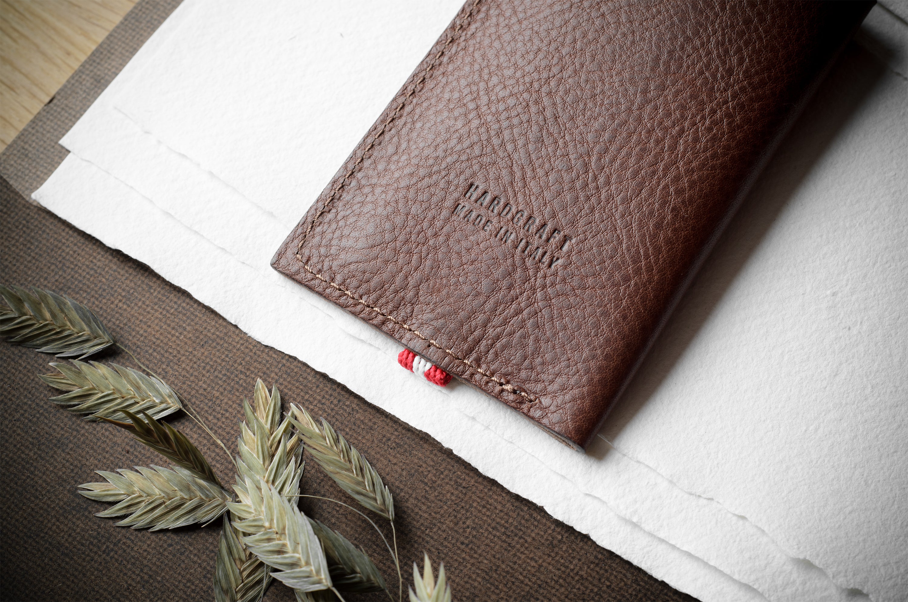 A brown textured leather notebook cover with 'HARDGRAFT MADE IN ITALY' debossed on the front, styled on a white sheet of paper with dried wheat stalks on a dark brown surface.