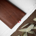 A brown textured leather card holder or wallet with 'HARDGRAFT MADE IN ITALY' embossed on the front, styled on a white paper background with a brown burlap section and dried oat stems.