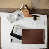 A brown leather wallet or card holder pouch lies on a white sheet of paper atop a clipboard, alongside cash, a credit card, and a gold-toned binder clip on a wood surface.